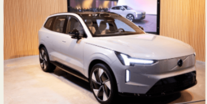 Bilden visar en grå elbil utställd i en modern lokal med en digital skärm i bakgrunden. Bilen har en elegant design med karaktäristiska linjer.
